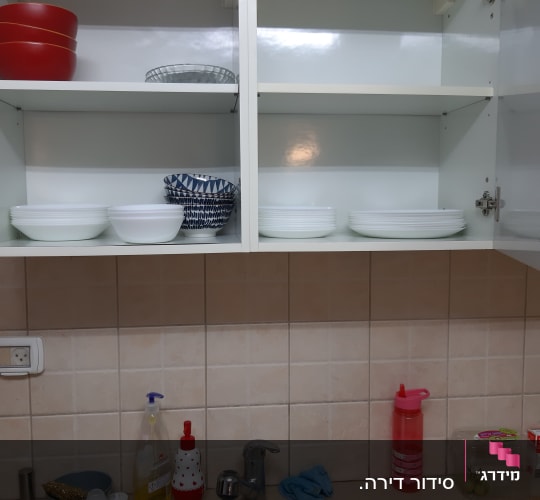 ארונות מטבח פתוחים עם כלים מסודרים, כיור עם כלים וחומרי ניקוי